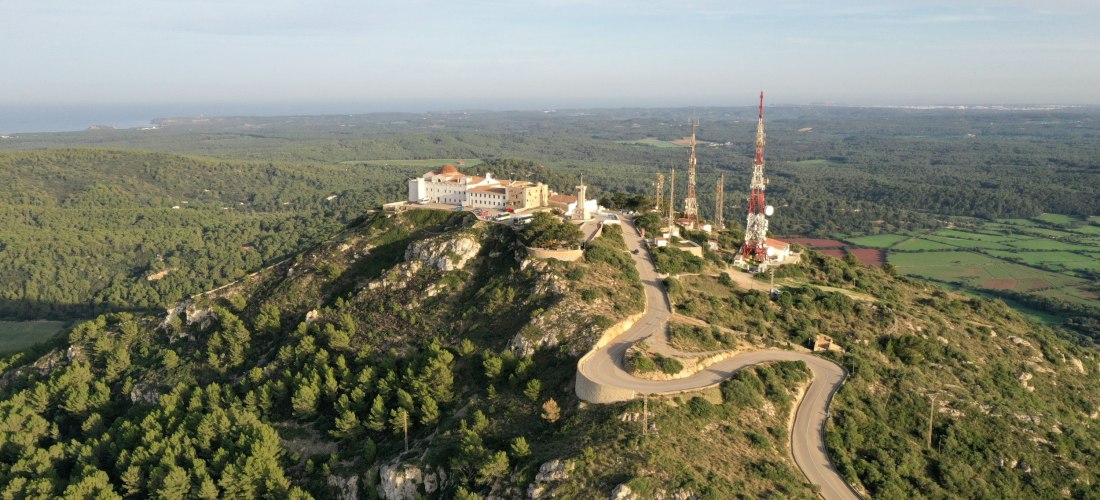 Luftaufnahme des Monte Toro, des höchsten Berges Menorcas, mit Klosteranlage, Sendemasten und einer kurvenreichen Straße, umgeben von grünen Wäldern und weiter Landschaft.