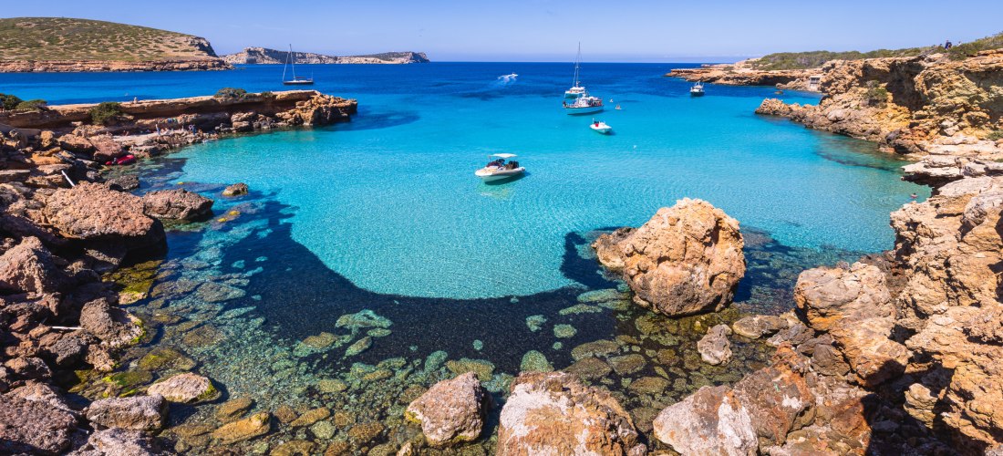 Felsige Bucht Cala Comte auf Ibiza mit kristallklarem, türkisfarbenem Wasser und mehreren Booten, die im ruhigen Meer ankern. Im Vordergrund ragen große Felsen ins Bild, dahinter die weite Küstenlandschaft.