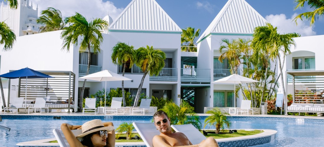 Ein Paar entspannt auf weißen Liegen im flachen Wasser eines Hotelpools. Die Sonne scheint, umgeben von Palmen und weißen Ferienhäusern mit hellblauen Fenstern. Die Atmosphäre wirkt ruhig und erholsam.