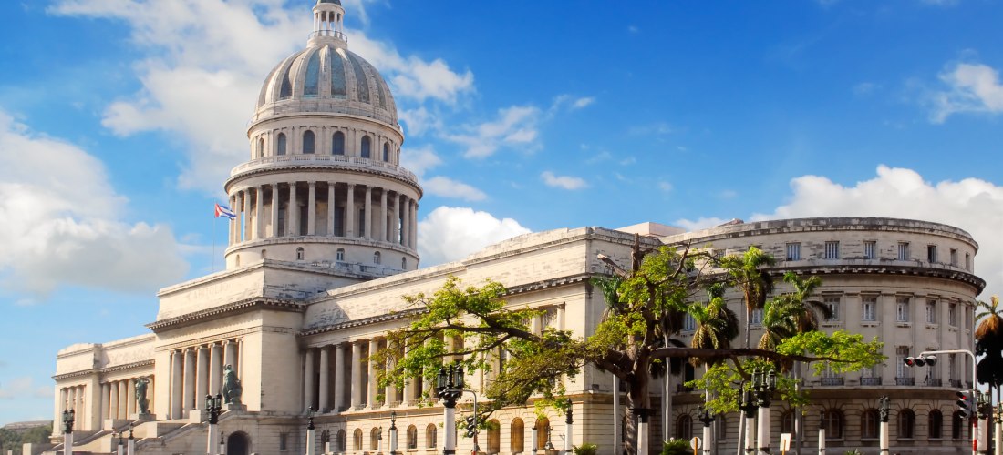 Das imposante Gebäude des El Capitolio in Havanna, Kuba. Vor dem klassizistischen Bauwerk mit Kuppel fahren farbenfrohe Oldtimer auf der Straße, umgeben von Palmen und blauen Himmel.