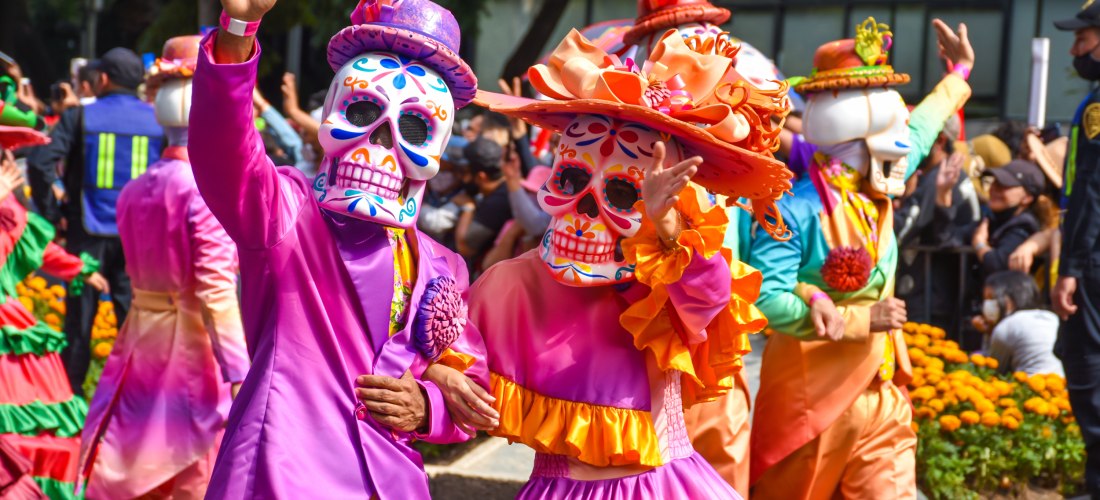 Eine farbenfrohe Parade zum Día de los Muertos (Tag der Toten) mit aufwendig geschmückten Teilnehmern in traditionellen Calavera-Masken und bunten, eleganten Kostümen. Die Feierlichkeiten ehren verstorbene Angehörige und sind ein wichtiger Bestandteil der