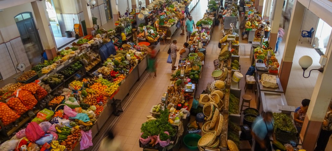 Blick von oben in eine überdachte Markthalle mit zahlreichen bunten Ständen voller frischer Früchte, Gemüse, Kräuter, Gewürze und lokaler Produkte. Menschen schlendern durch die Gänge zwischen den Verkaufsständen.