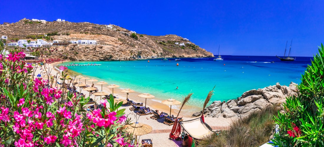 Blick auf den Beach auf Mykonos mit türkisblauem Meer, Strohdach-Sonnenschirmen und Segelbooten in der Bucht