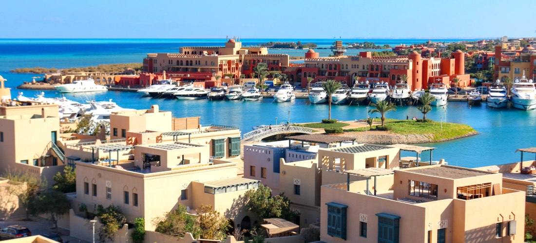 Blick auf den Yachthafen von El Gouna, Ägypten. Im Vordergrund befinden sich moderne, helle Wohnhäuser im nubisch-arabischen Stil. Dahinter liegen zahlreiche weiße Luxusyachten vor dem orientalisch anmutenden Gebäudeensemble der Abu Tig Marina. Das tiefbl