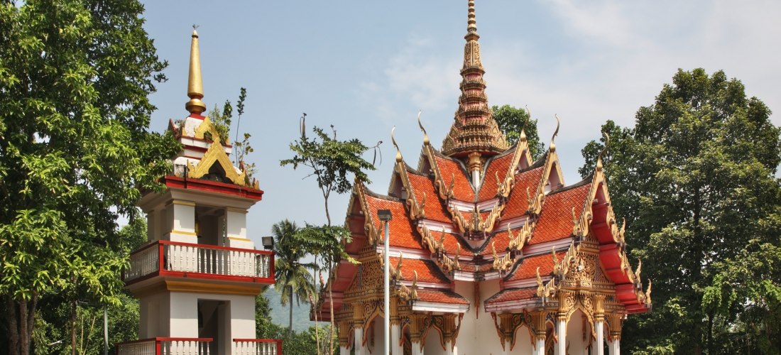 Ein traditioneller thailändischer Tempel auf Phuket, mit kunstvoll verziertem, spitz zulaufendem Dach in Rot und Gold. Daneben ein kleiner weiß-roter Glockenturm. Der Tempel ist von tropischen Bäumen umgeben und liegt in einer ruhigen Umgebung. ( Wat Ket 
