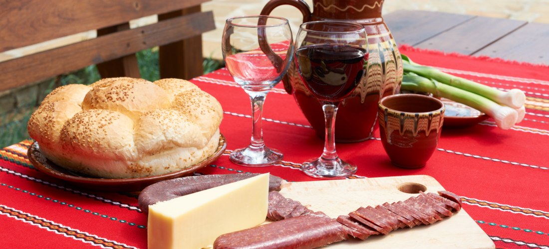 Ein gedeckter Holztisch im Freien mit einer traditionellen roten Tischdecke. Darauf ein großer runder Laib Sesambrot, zwei Weingläser – eines mit Rotwein gefüllt, das andere leer –, eine braune Tonkaraffe, Frühlingszwiebeln und ein Holzbrett mit Käsestück