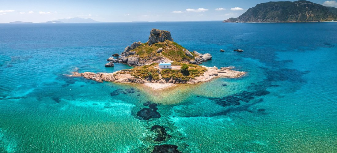 Drohnenbild der kleinen Insel Kastri vor Kos mit einer weiß-blauen Kapelle, umgeben von türkisfarbenem Wasser. Die Insel ist von Felsen umgeben, im Hintergrund liegt die grüne Küste der Halbinsel.