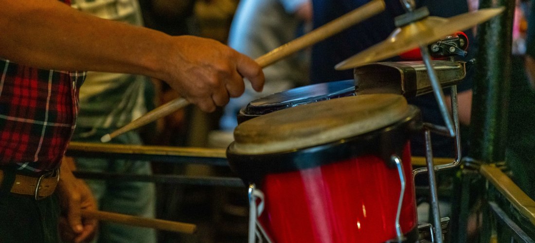 Ein Musiker spielt mit Drumsticks auf roten Bongos und einem kleinen Schlagzeug in einer lebhaften Umgebung