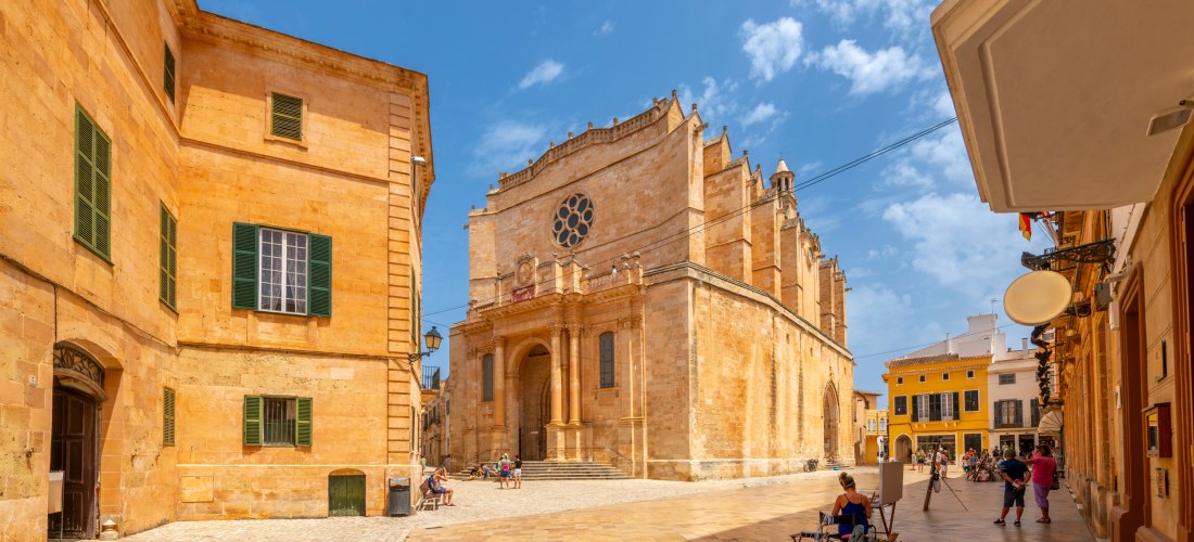 Helle Sandsteinfassaden umgeben einen sonnigen Platz in Ciutadella auf Menorca. Im Zentrum steht die beeindruckende Kathedrale Santa Maria mit gotischer Fassade und einem großen Rosettenfenster. Auf dem Platz sitzen Menschen entspannt im Schatten, einige 