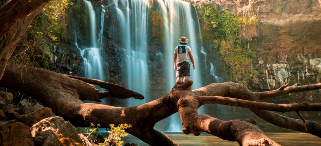 Ein Mann steht auf einem mächtigen umgestürzten Baumstamm, der über ein ruhiges Gewässer vor einem breiten Wasserfall ragt. Der Wasserfall stürzt in mehreren Kaskaden von einer Felskante herab, umgeben von dichtem Grün. Die Szene wirkt wild, abenteuerlich