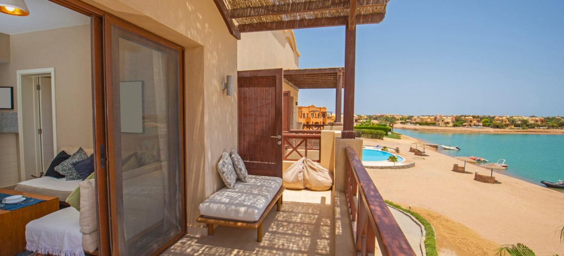 Blick von einem überdachten Balkon einer Ferienunterkunft direkt an einer Lagune in El Gouna, Ägypten. Auf dem Balkon befinden sich eine gepolsterte Sitzbank und Sitzsäcke. Rechts im Bild erstreckt sich ein ruhiger Sandstrand mit Blick auf türkisblaues Wa