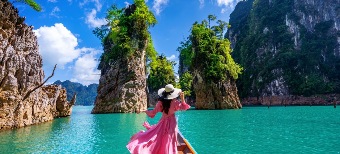 Frau in pinkem Kleid auf Langboot zwischen tropischen Felsen im türkisfarbenen Wasser unter blauem Himmel.