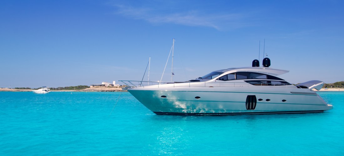 Moderne weiße Motoryacht liegt auf hellblauem, glasklarem Wasser vor Anker. Im Hintergrund ist eine flache Küstenlinie mit vereinzelten Gebäuden zu sehen. Der Himmel ist wolkenlos und strahlend blau.