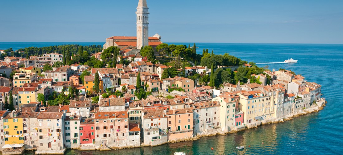 Blick auf die Altstadt von Rovinj in Kroatien. Bunte Häuser stehen dicht an dicht am Wasser, dahinter erhebt sich die barocke Kirche der Heiligen Euphemia mit ihrem hohen Glockenturm. Im Hintergrund das glitzernde Blau der Adria.