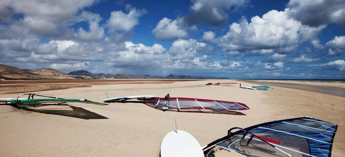 Mehrere Windsurf-Boards mit Segeln liegen am sandigen Strand unter einem teils bewölkten Himmel. Im Hintergrund Berge und Weite der Küste