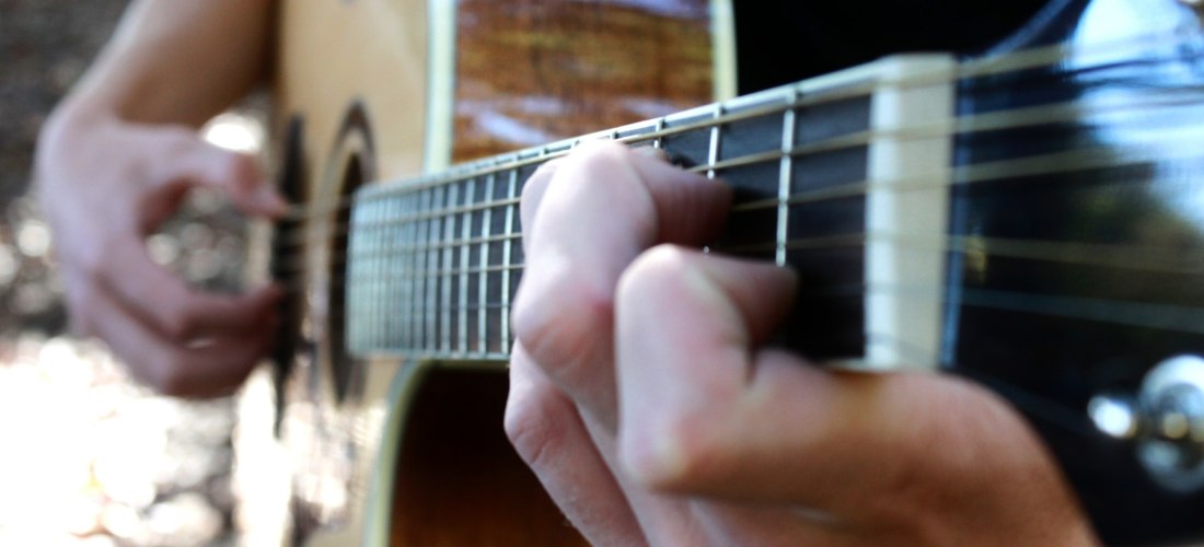 Nahaufnahme einer Hand, die auf einer akustischen Gitarre spielt. Die Finger drücken die Saiten auf das Griffbrett, während die andere Hand die Saiten anschlägt.