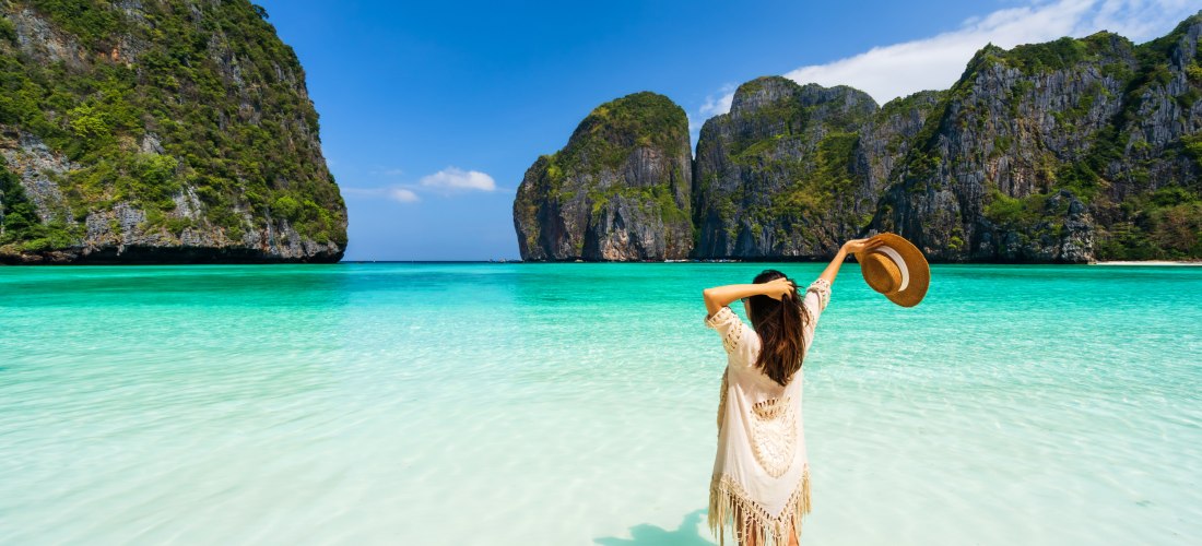 Das Bild zeigt eine Frau, die barfuß im flachen, kristallklaren Wasser der berühmten Maya Bay auf Ko Phi Phi Leh steht. Sie trägt einen cremefarbenen, offenen Strandponcho mit Fransen und hält ihren Sonnenhut in einer Hand, während sie mit der anderen dur