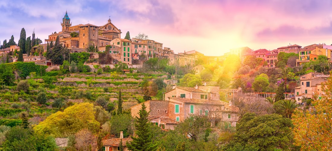 Malerisches Bergdorf auf Mallorca bei Sonnenuntergang. Die historischen Steinhäuser mit grünen Fensterläden schmiegen sich an die terrassierten Hänge, umgeben von üppigem Grün, Zypressen und anderen Bäumen. Im oberen Teil des Bildes ist eine Kirche mit ei