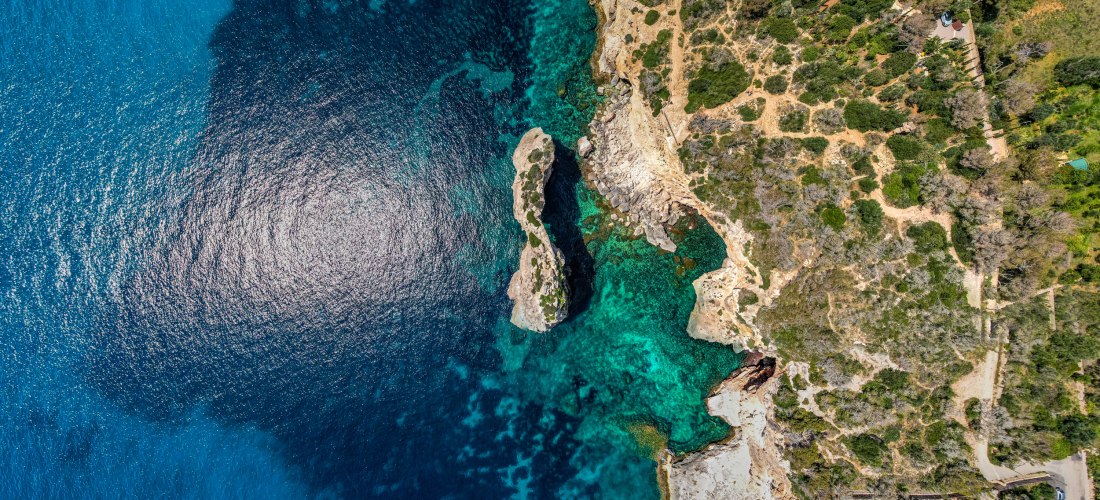 Luftaufnahme der zerklüfteten Küste von Mallorca mit türkisblauem Meer, Felsen und einzelnen Häusern am Rand der Klippen