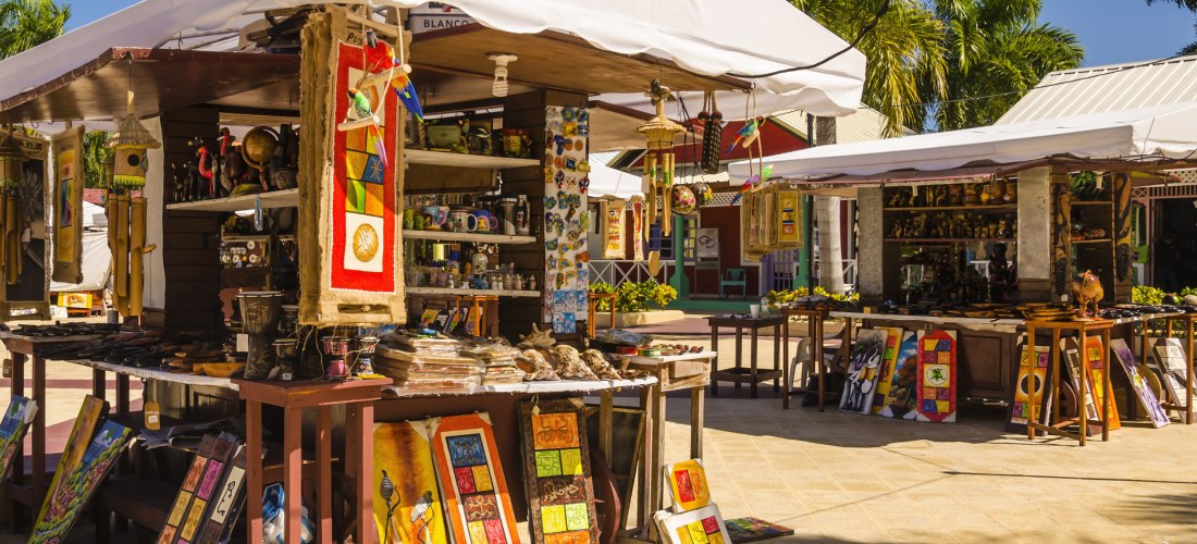 Fischerdorf Oistins: Bunte Marktstände auf Barbados mit handgemachten Kunstwerken, Wandbildern, Töpferwaren und Souvenirs. Die Stände sind mit weißen Sonnendächern überspannt, Palmen im Hintergrund
