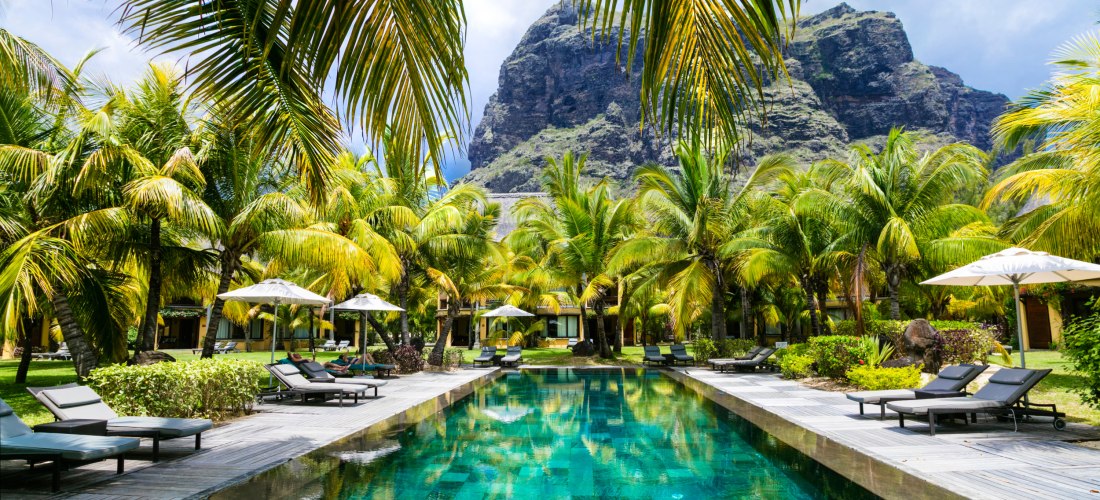 Dieses Bild zeigt eine traumhafte Urlaubsidylle: Ein großes, elegantes Resort mit einem türkisfarbenen Infinity-Pool, der von hohen, sattgrünen Palmen eingerahmt wird. Schwarze Sonnenliegen mit weißen Sonnenschirmen laden zum Entspannen ein. Im Hintergrun