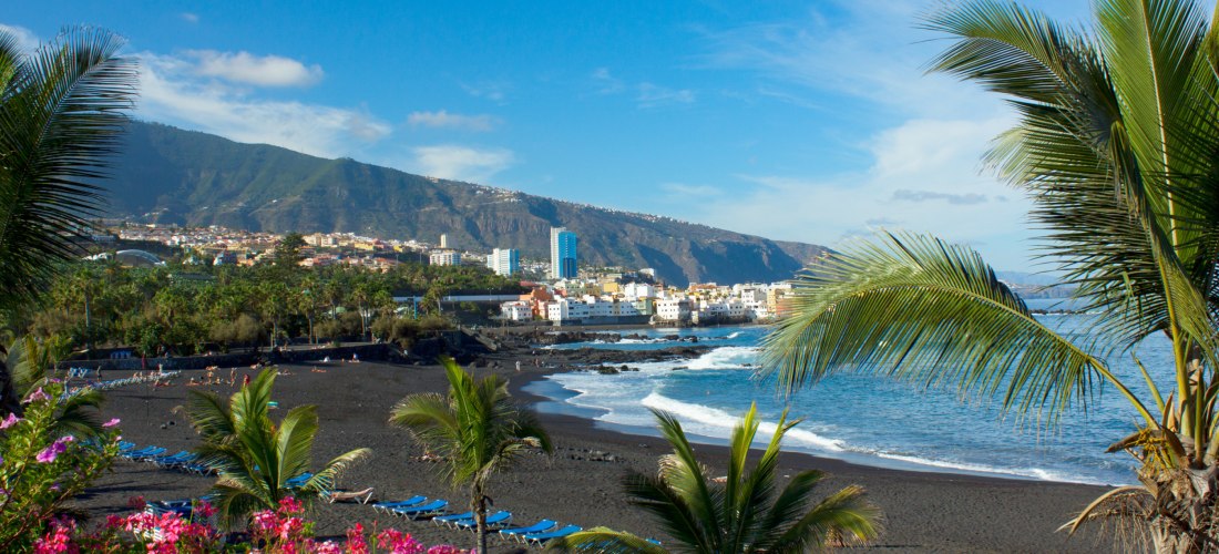 Playa Jardín in Puerto de la Cruz, Teneriffa: schwarzer Sandstrand mit Palmen, Blumen, Liegen und Blick auf die Stadt und Berge im Hintergrund.