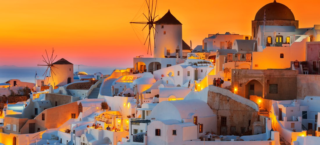 Romantische Abendstimmung über Oia auf Santorini: Die weiß getünchten Häuser mit blauen Kuppeln leuchten im goldenen Licht des Sonnenuntergangs. Im Zentrum eine ikonische Windmühle. Der Himmel ist orangefarben und taucht die gesamte Szenerie in warmes Lic