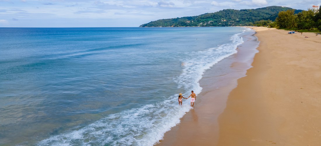 Paar spaziert Hand in Hand am Karon Beach auf Phuket entlang des feinen Sandstrandes mit türkisblauem Meer