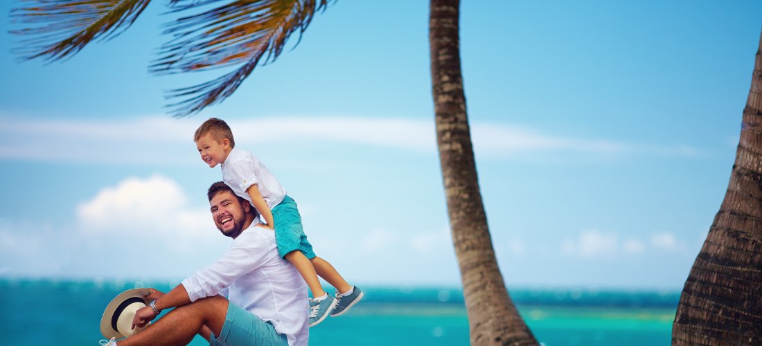 Lachender Vater in Sommerkleidung mit seinem Sohn auf den Schultern, am tropischen Strand mit Palmen und türkisblauem Meer