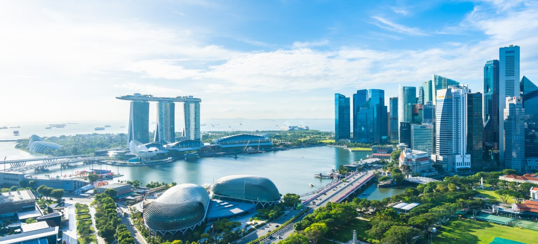 Panoramablick auf die moderne Skyline von Singapur bei Tageslicht. Links ist das ikonische Marina Bay Sands Hotel mit seinen drei Türmen und der verbindenden Dachterrasse zu sehen. Davor liegt die Marina Bay mit ruhigem Wasser, umgeben von futuristischen 