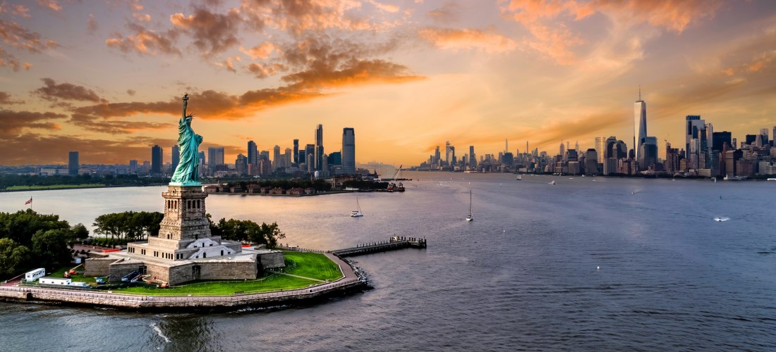 Die Freiheitsstatue auf Liberty Island vor der Skyline von New York City bei Sonnenuntergang. Der Himmel ist orange gefärbt, Boote fahren auf dem Wasser, und das One World Trade Center ragt hoch über die restlichen Gebäude hinaus.