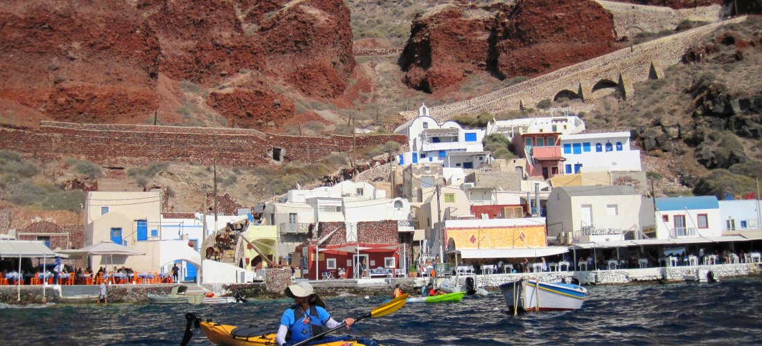 Ein gelbes Kajak mit einer Frau im blauen Oberteil auf dem Wasser vor Ammoudi Bay auf Santorini. Im Hintergrund sind bunte Häuser direkt an der Küste zu sehen, die sich an rote Felsen schmiegen. Darüber liegen die weißen Gebäude von Oia