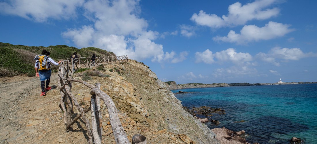 Mehrere Wanderer gehen einen schmalen Küstenpfad auf Menorca entlang, gesichert mit einem rustikalen Holzgeländer. Der Weg führt über eine felsige Anhöhe mit Blick auf das tiefblaue Meer. Am Horizont ist ein Leuchtturm zu sehen. Der Himmel ist mit weißen 