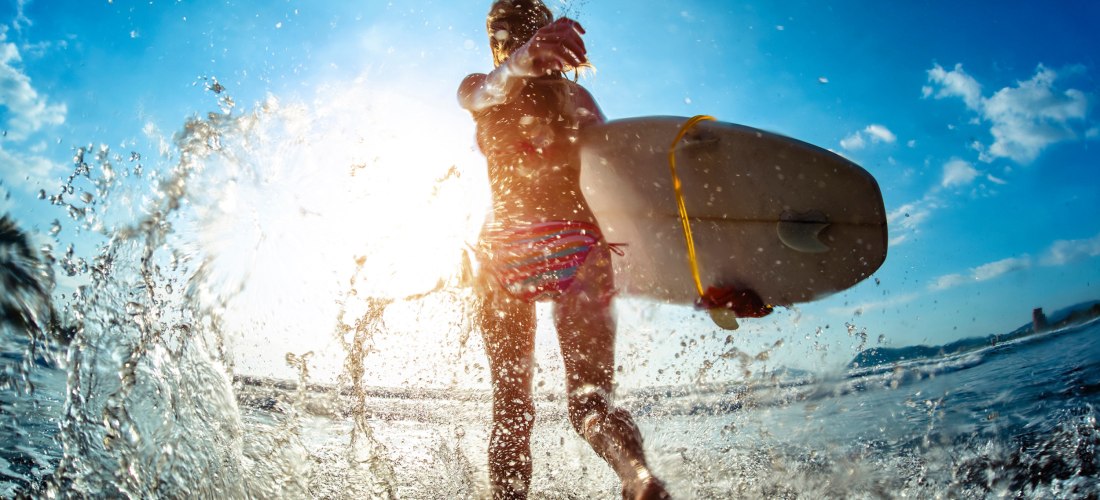 Surferin mit Surfbrett läuft durch spritzendes Wasser bei Sonnenlicht, mit blauem Himmel im Hintergrund