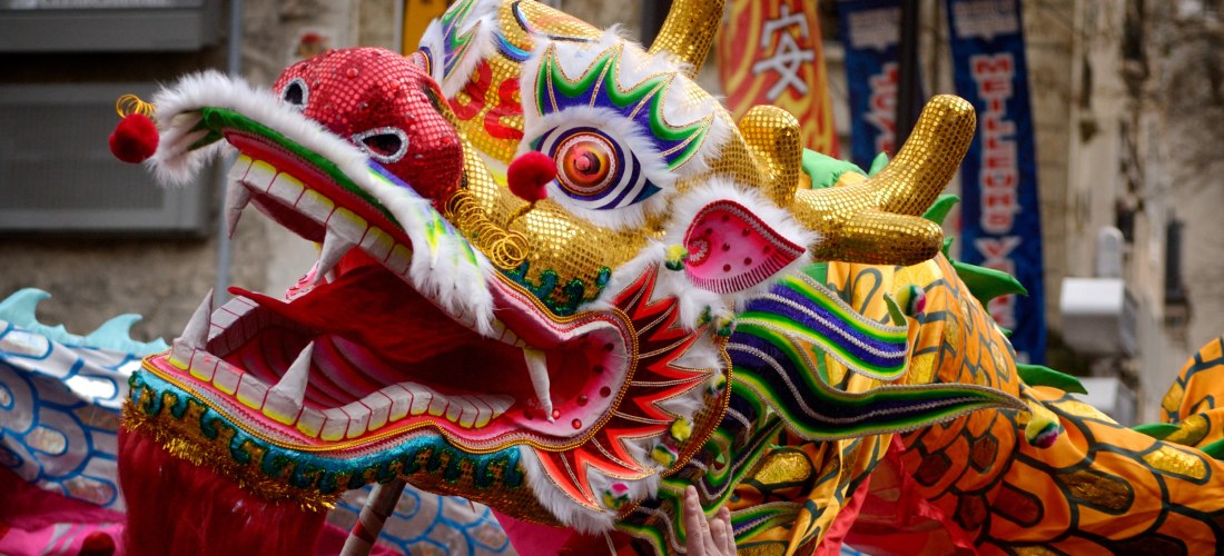 eindrucksvolle Darstellung eines chinesischen Drachentanzes während einer Parade. Der Drache ist farbenprächtig gestaltet mit einem goldglänzenden Kopf, roten Augen, aufwendigen Mustern und goldenen Hörnern. Sein offenes Maul mit scharfen Zähnen und einer