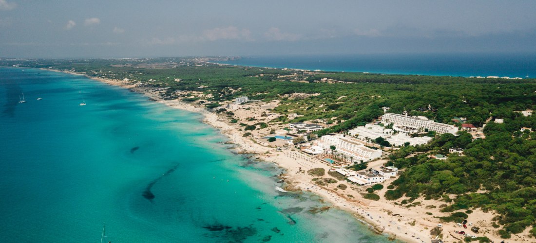 Luftaufnahme eines langen Strandes mit klarem, türkisfarbenem Wasser und einem Hotelkomplex im Grünen auf Formentera.