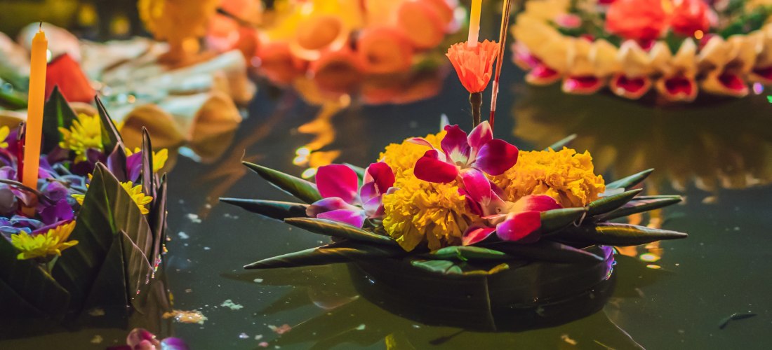 Mehrere bunt geschmückte Krathongs aus Blumen und Bananenblättern mit brennenden Kerzen schwimmen auf dunklem Wasser. Das Licht der Kerzen spiegelt sich im Wasser, was eine festliche und spirituelle Atmosphäre erzeugt. Im Vordergrund ein Krathong mit gelb