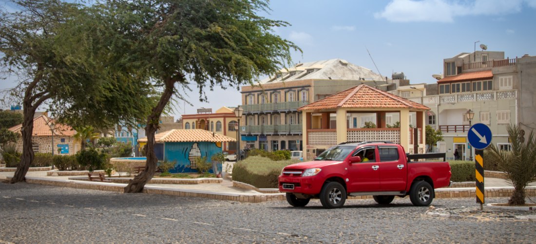 Straßenszene auf den Kapverden mit einem roten Pickup, der auf einer gepflasterten Straße an pastellfarbenen Häusern vorbeifährt. Im Hintergrund stehen moderne und traditionelle Gebäude, sowie ein bemaltes Wandbild mit maritimem Motiv.