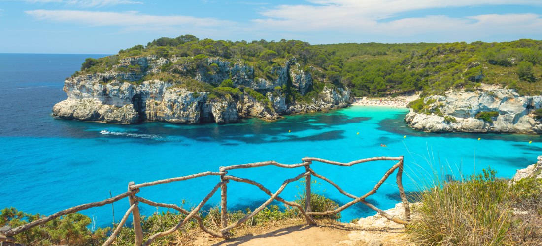 Atemberaubender Aussichtspunkt mit rustikalem Holzgeländer über einer türkisblauen Bucht auf Menorca. Im Hintergrund liegen helle Kalkfelsen, grüne Pinienwälder und ein kleiner Sandstrand mit Badenden. Das Wasser leuchtet in kräftigem Blau, ein Boot hinte