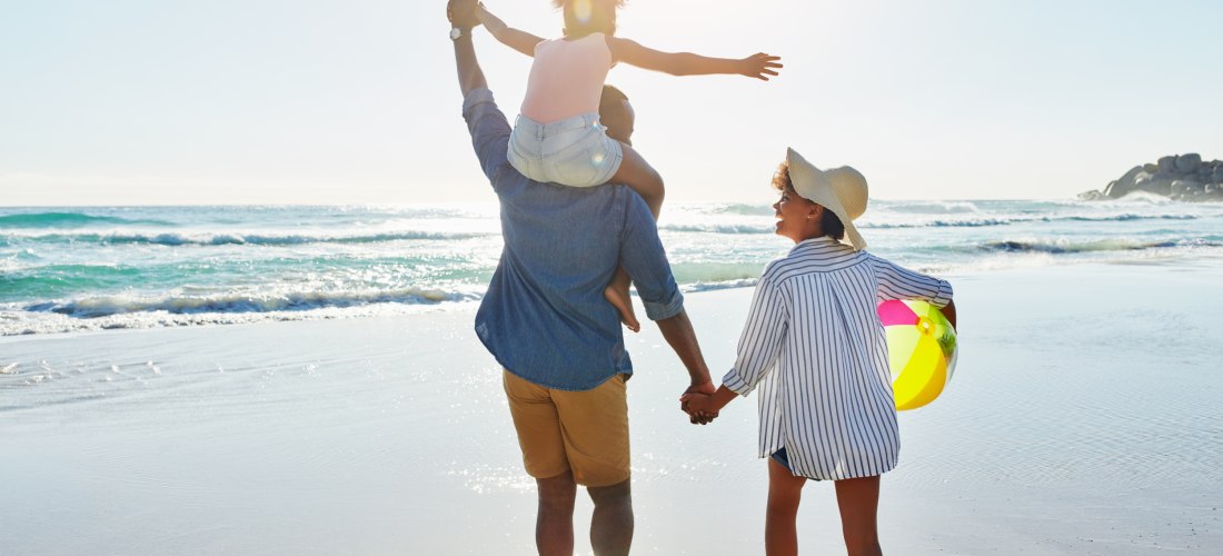 Eine fröhliche Familie spaziert barfuß am sonnigen Strand entlang. Der Vater trägt ein Kind auf den Schultern, das lachend die Arme ausbreitet. Die Mutter hält die Hand des Vaters und trägt einen bunten Wasserball sowie einen Sonnenhut. Im Hintergrund sch
