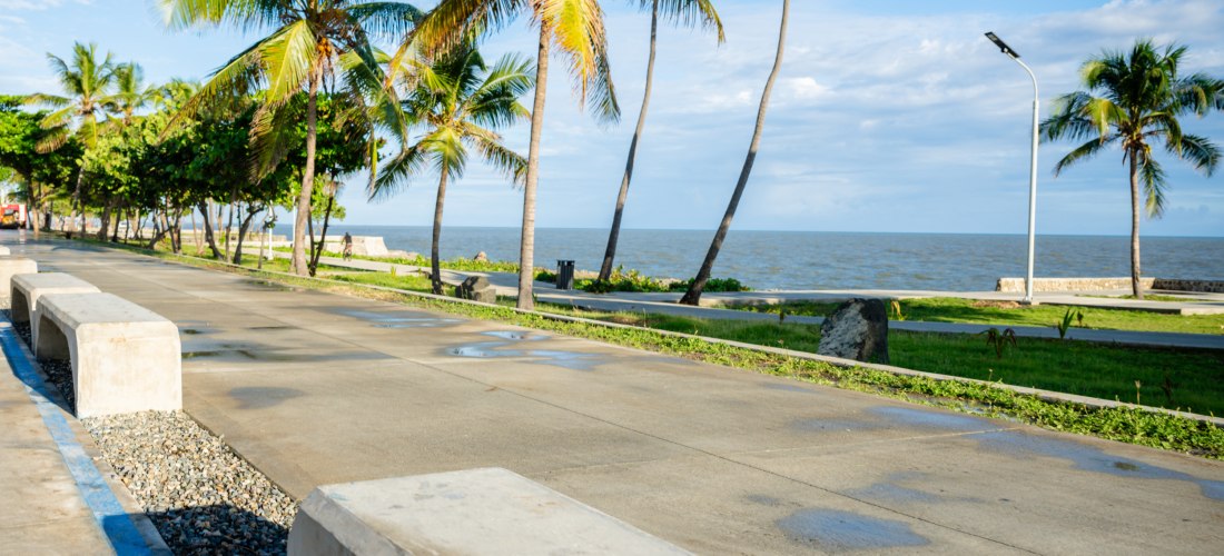 Ruhige Promenade mit Betonbänken und Palmen direkt am Meer unter einem klaren Himmel, mit vereinzelten Wasserpfützen auf dem Weg