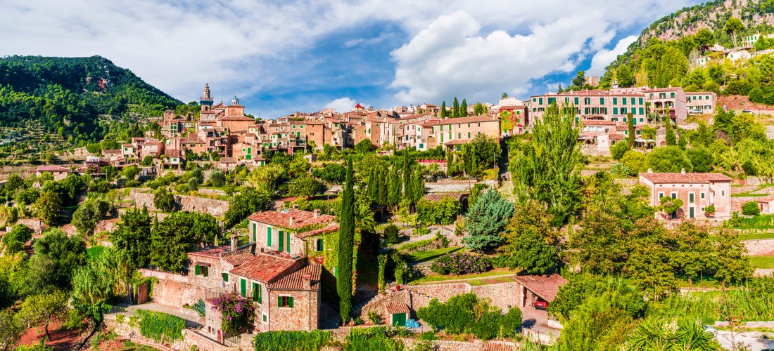 Ein malerischer Blick auf ein mediterranes Dorf auf Mallorca, das sich an einen Hang schmiegt. Die Häuser bestehen aus Naturstein mit grünen Fensterläden und sind von Zypressen, Pinien und anderen mediterranen Pflanzen umgeben. Der Hintergrund zeigt einen