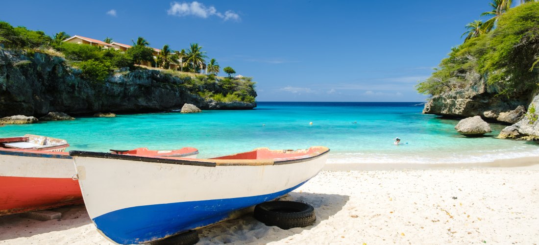 Zwei kleine Fischerboote stehen auf dem hellen Sandstrand einer türkisblauen Bucht. Die Boote sind weiß lackiert mit roten und blauen Akzenten. Im Hintergrund ragen grüne Felsen ins Meer, darüber Palmen und gelb gestrichene Häuser mit roten Dächern. Das r
