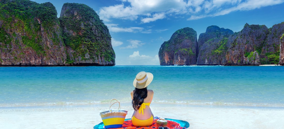 Frau sitzt am weißen Sandstrand der Maya Bay auf den Phi Phi Inseln bei Phuket, Thailand, mit Blick auf türkisfarbenes Meer und Kalksteinfelsen.