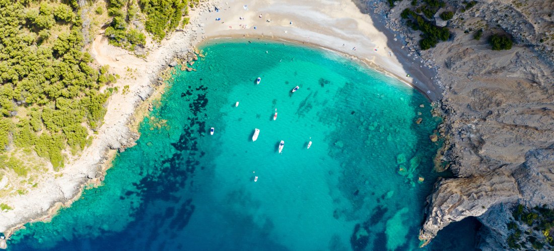 Luftaufnahme von Platja des Coll Baix, einer abgelegenen Badebucht im Norden Mallorcas. Die sichelförmige Bucht ist von steilen Felsen eingerahmt und nur zu Fuß oder mit dem Boot erreichbar. Der Strand besteht aus hellem Kies und Sand, das Wasser leuchtet
