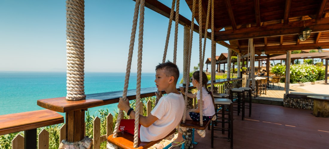  Pause mit Aussicht auf Korfu: Zwei Kinder genießen den Blick aufs Meer von Schaukeln aus, die an einer gemütlichen Holzterrasse befestigt sind. Das Café ist liebevoll mit Stein und Holz gestaltet. Der Ausblick reicht über das türkisfarbene Wasser bis zum