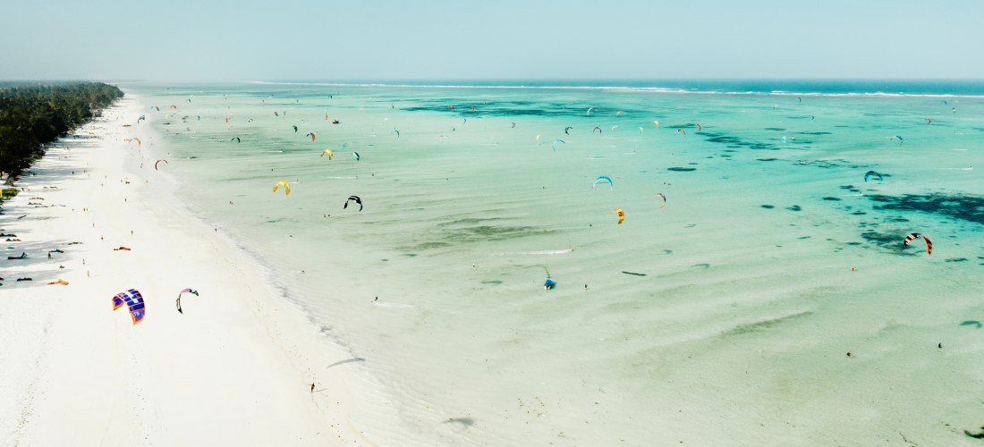 Weißer Sandstrand auf Sansibar mit türkisblauem Meer voller Kitesurfer – beliebter Spot für Wassersport und Aktivitäten im Urlaub in Tansania