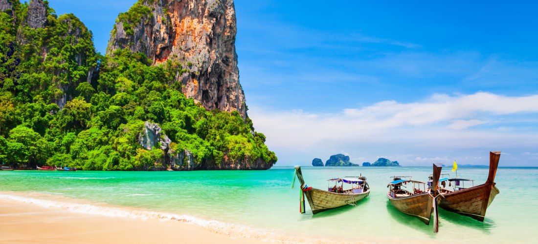 Das Bild zeigt einen feinsandigen, tropischen Strand mit flach auslaufendem türkisblauem Meer. Im Vordergrund liegen drei typische thailändische Longtailboote im Wasser, mit bunten Tüchern und einem gelben Fähnchen geschmückt. Dahinter erhebt sich eine im