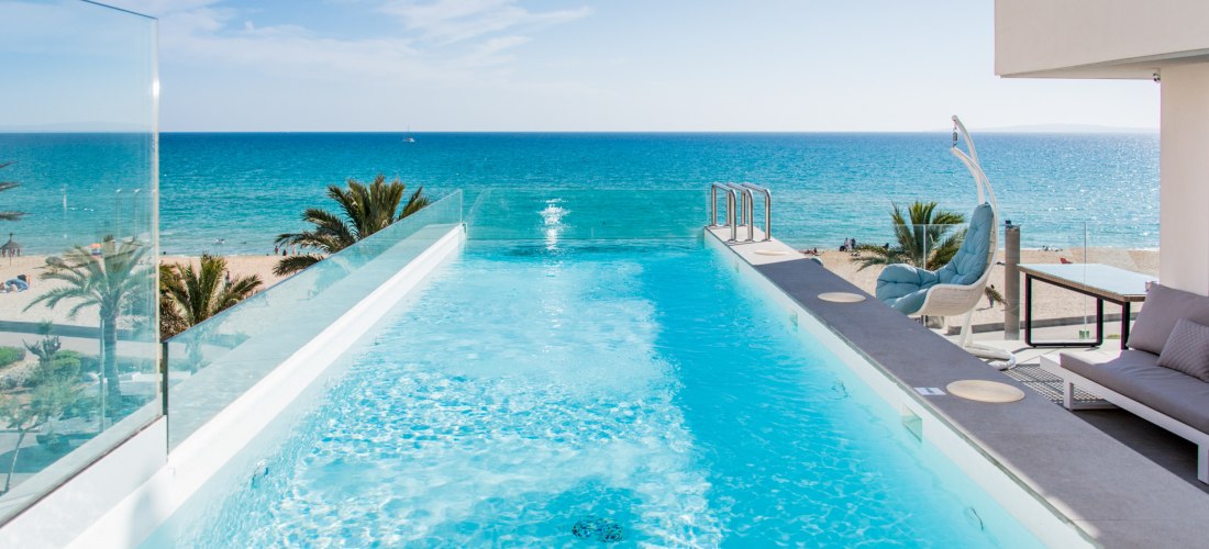 Infinity-Pool mit klarem, hellblauem Wasser auf einer Dachterrasse. Dahinter ein Sandstrand, Palmen und das türkisblaue Meer unter wolkenlosem Himmel.
