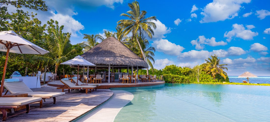 Dieses Bild zeigt eine exklusive Urlaubsoase mit einem atemberaubenden Infinity-Pool, der nahtlos in den Horizont übergeht. Die Poollandschaft ist umgeben von eleganten Holzliegen mit weißen Polstern, die unter großen Sonnenschirmen für Schatten sorgen. I